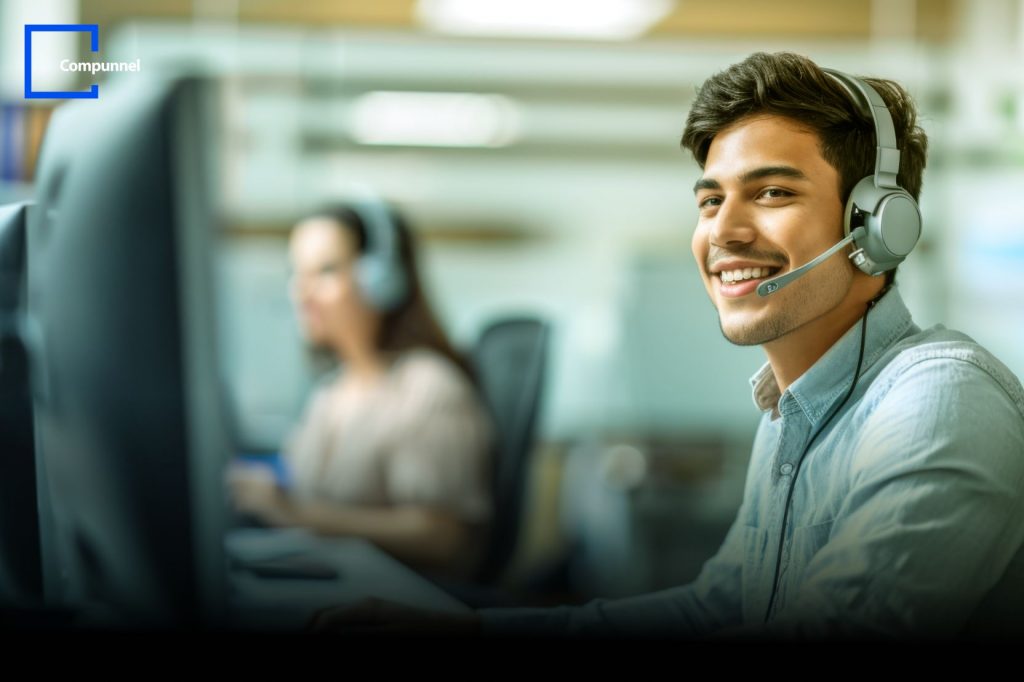 Smiling customer service representative wearing a headset, sitting at a desk in a call center, with Compunnel logo in the top left corner.